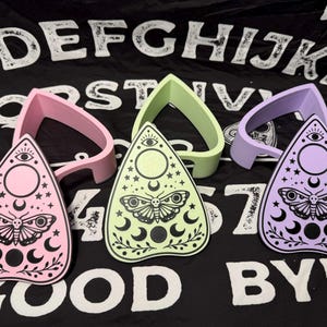 May include: Three planchette boards in pink, green, and purple, each with a matching planchette guide. The boards feature a moth, moon, and star design. The background is a black cloth with white letters spelling out "GOOD BYE".