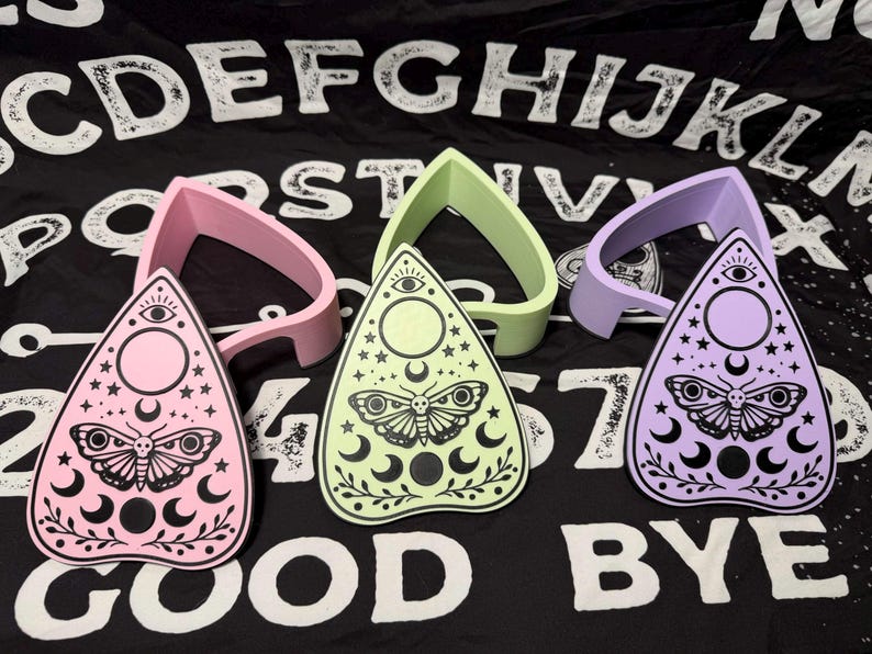 May include: Three planchette boards in pink, green, and purple, each with a matching planchette guide. The boards feature a moth, moon, and star design. The background is a black cloth with white letters spelling out "GOOD BYE".
