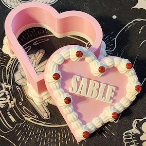 May include: A pink heart-shaped cookie cutter next to a pink heart-shaped object with the name "SABLE" in white letters. The heart-shaped object has white frosting and red cherry-like decorations around the edge. The background is black with white illustrations.
