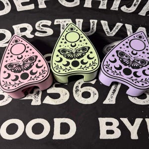 Può includere: Tre scatole pastello a forma di planchette. Ogni scatola presenta un motivo a falena e mezzaluna con stelle e un simbolo dell'occhio. Le scatole sono rosa, verdi e viola, su uno sfondo a motivi bianchi e neri.