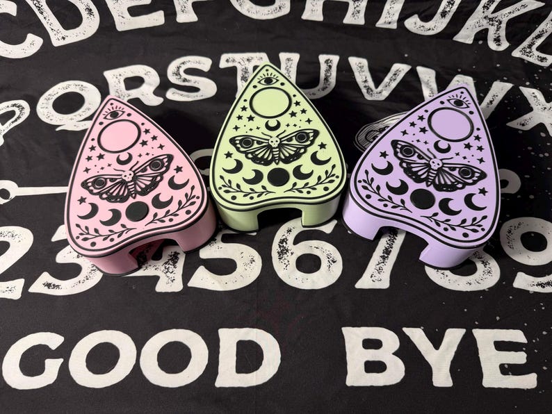 May include: Three pastel-colored planchette-shaped boxes. Each box features a moth and crescent moon design with stars and an eye symbol. The boxes are pink, green, and purple, set against a black and white patterned background.