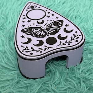 May include: A lavender-colored, decorative planchette with a black moth and celestial design. The planchette features a black border and is set against a teal, fuzzy background. The design includes moons, stars, and an eye symbol.