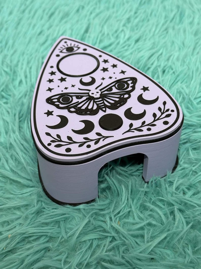 May include: A lavender-colored, decorative planchette with a black moth and celestial design. The planchette features a black border and is set against a teal, fuzzy background. The design includes moons, stars, and an eye symbol.