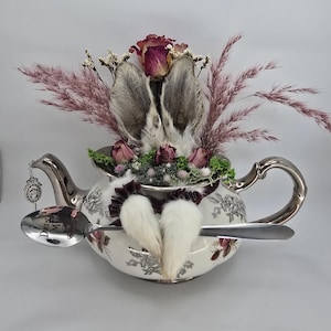 May include: A decorative teapot with a floral design, featuring rabbit ears and a tail, along with dried flowers and a spoon. The spoon has the words "DRINK ME" engraved on it. The teapot is white with silver accents.