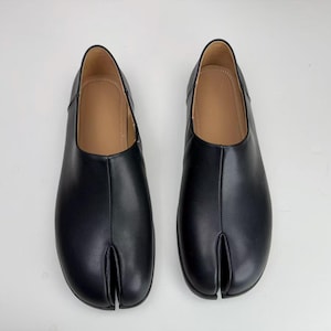 Op de afbeelding: Een paar zwarte leren schoenen met split-teen. De schoenen hebben een uniek ontwerp met een splitsing tussen de grote teen en de andere tenen. De binnenkant is lichtbruin van kleur. De schoenen worden op een wit oppervlak gepresenteerd.