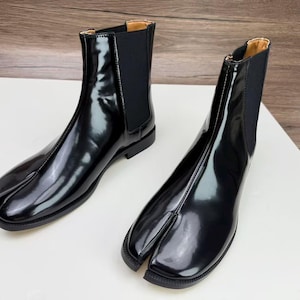 May include: Pair of black leather ankle boots with a split-toe design. The boots have a glossy finish, elastic side panels, and a low heel. The boots are displayed on a white surface.