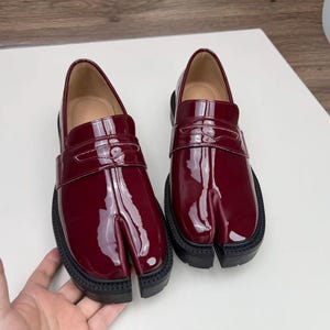 May include: A pair of burgundy leather split-toe shoes with a glossy finish. The shoes have a unique design with a separate section for the big toe and a thick black sole. The shoes are displayed on a white surface.