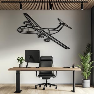 May include: A black metal wall art piece shaped like a large cargo airplane with four engines, mounted on a plain wall above a wooden desk, a black office chair, a computer monitor, a small potted plant, and a tall green floor plant.