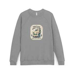May include: A heather gray crewneck sweatshirt featuring a central graphic of a beige cat sitting in a teal armchair. The text reads, "This is my quiet corner. Only cats are allowed," framed within a cream-colored rounded rectangular border.