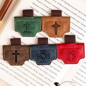 May include: Leather bookmarks in green, brown, dark brown, blue, and red, each with a cross design. The bookmarks have a loop for inserting into a book. A pen rests on the open book.