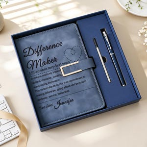 May include: A blue notebook with a gold-tone metal clasp and the text Difference Maker, a definition, and the name Jennifer, presented in a blue gift box alongside a matching blue and silver pen and a thin metal stylus on a light surface.
