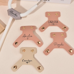 May include: A collection of personalized stethoscope tags in various colors, including pink and beige. Each tag features a name, such as "Amelia" or "Carolyn", and decorative elements. The tags are designed to attach to a stethoscope.