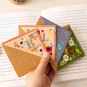 Puede incluir: Una mano sostiene cuatro marcapáginas de fieltro bordado con diseños florales e iniciales. Los marcapáginas son de color beige, gris y verde, con costuras rojas y azules. Están sobre un libro abierto.