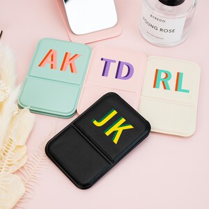 May include: Four rectangular folding mirrors in mint green, light pink, cream, and black. Each mirror features two-letter monograms: AK in orange and red, TD in purple, RL in red and teal, and JK in yellow and green. A Byredo perfume bottle is nearby.