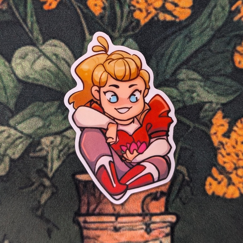 Pode incluir: Um adesivo com um personagem de desenho animado com cabelo loiro em rabo de cavalo, vestindo uma roupa vermelha e branca e segurando um objeto rosa em forma de cora&ccedil;&atilde;o. O adesivo tem uma borda branca e est&aacute; sobre um fundo floral verde escuro.