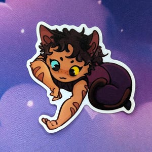 Pode incluir: Um autocolante com uma personagem de desenho animado semelhante a um gato com pelo castanho, uma roupa roxa e um contorno branco. A personagem tem um olho azul e outro amarelo e est&aacute; deitada com o bra&ccedil;o esticado.