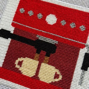 May include: Needlepoint design of a red espresso machine with a black portafilter and two beige mugs. The machine has silver accents and is set against a white background. The design is a close-up view of the coffee maker.