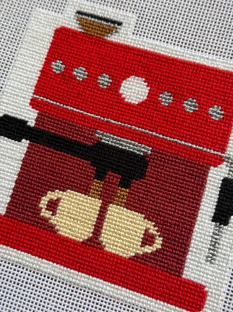 May include: Needlepoint design of a red espresso machine with a black portafilter and two beige mugs. The machine has silver accents and is set against a white background. The design is a close-up view of the coffee maker.