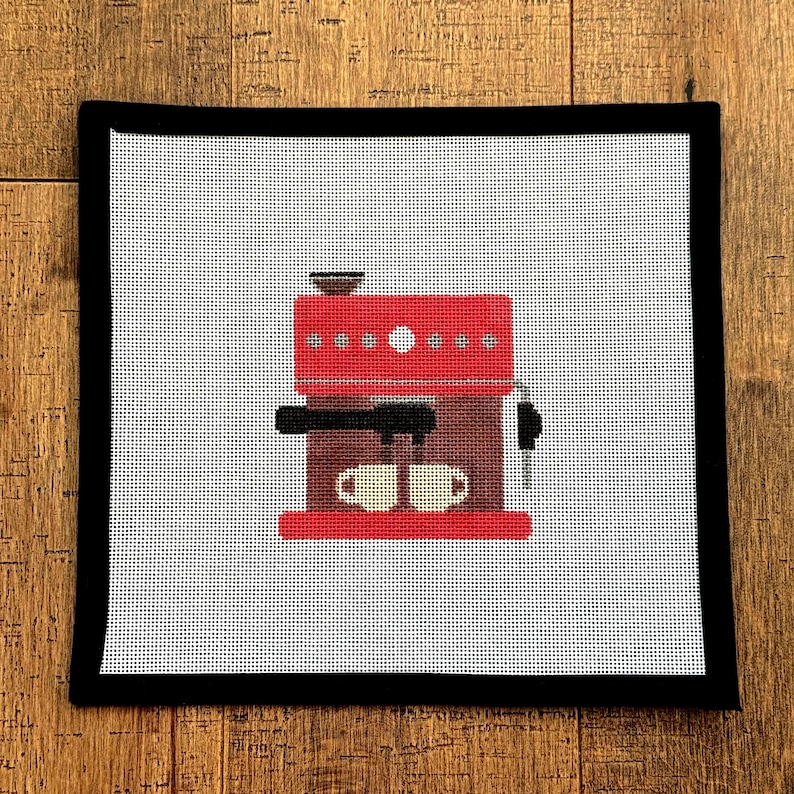 May include: A needlepoint canvas featuring a red espresso machine with two cups. The machine has a dark brown base and black details. The canvas is bordered by a black fabric.