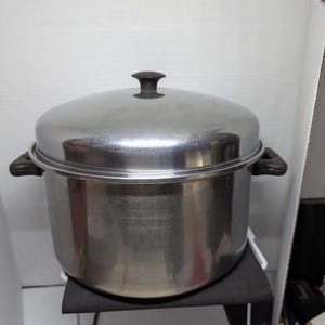 May include: A stainless steel steamer pot with a lid and two side handles. The pot is sitting on a black surface. The lid has a dark brown handle.