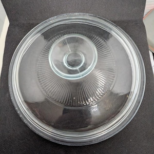 Op de afbeelding: Een heldere glazen deksel met een cirkelvormig ontwerp. De deksel heeft een centrale knop en concentrische ringen met radiale lijnen. De deksel is ontworpen voor een pot of pan.