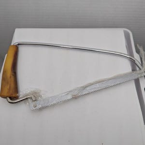 May include: A metal butcher's saw with a tan handle. The saw blade is wrapped in clear plastic. The saw has a curved metal frame and a toothed blade, designed for cutting meat or bone.