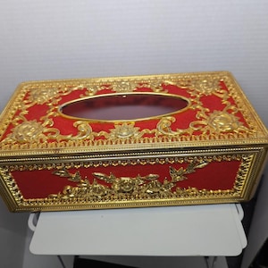 May include: A rectangular tissue box with a red velvet exterior and ornate gold-coloured metal detailing. The box features a central oval opening and floral and scrollwork designs. The box is approximately 23 cm long, 13 cm wide, and 8 cm tall.