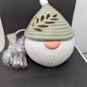 May include: A ceramic gnome-shaped electric wax warmer. The warmer has a white beard, a pink nose, and a green hat with leaf-shaped cutouts. A white bulb tops the hat. A power cord with a plug is attached.