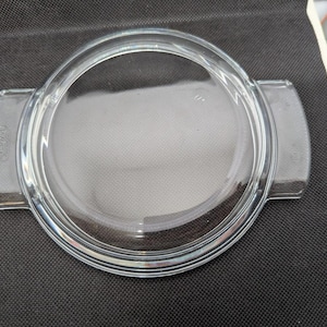 May include: A clear glass Pyrex lid with a circular design and two side handles. The lid is transparent, allowing a view of the surface beneath. The handles are rectangular and integrated into the lid's design. The overall shape is round.