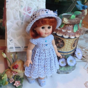 May include: A doll wearing a lavender crocheted dress and a white crocheted hat with a blue ribbon. The doll has red hair and blue eyes.