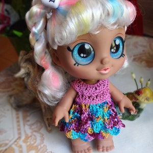 Puede incluir: Una muñeca con el pelo largo y blanco, peinado en trenzas con mechones de colores. La muñeca tiene grandes ojos azules y lleva un vestido de ganchillo rosa y morado.