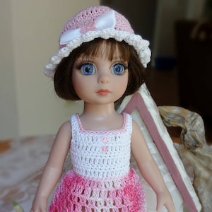 Peut inclure: Une poupée portant une robe et un chapeau en crochet rose et blanc. La poupée a les yeux bleus et les cheveux bruns.
