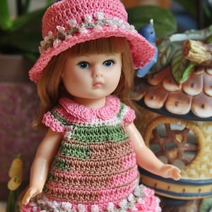 以下が含まれることがあります： ピンクとグリーンのドレスを着た、ピンクのクロシェハットをかぶった、クロシェのドール。ドールは茶色の髪と青い目です。