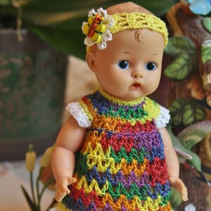 May include: A doll wearing a rainbow crochet dress with a white crocheted trim and a yellow crocheted headband with a butterfly accent.