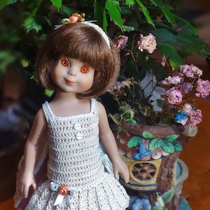May include: A doll wearing a crocheted dress with a flower accent. The doll has brown hair and blue eyes. The doll is standing in front of a miniature treehouse.