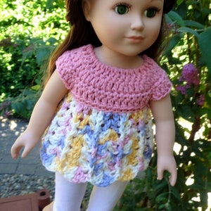 May include: A doll wearing a pink and white crocheted dress with a scalloped hem. The doll has long brown hair and green eyes.