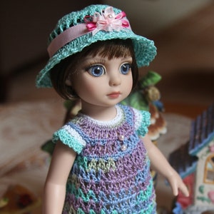 May include: A doll wearing a crocheted dress in a light purple, blue and green colour scheme. The dress has a white crocheted trim and a pink ribbon around the waist. The doll is also wearing a light blue crocheted hat with a pink flower.