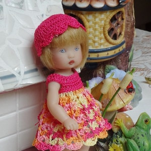 May include: A small doll wearing a pink crocheted dress and hat. The doll has blonde hair and blue eyes. The doll is standing on a white surface with a green frog figurine and a mushroom figurine in the background.