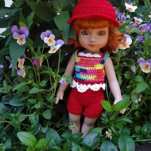 Może przedstawiać: Lalka w czerwonym, szydełkowym kapeluszu, tęczowym, szydełkowym topie z białym wykończeniem i czerwonymi, szydełkowymi spodenkami. Lalka stoi w ogrodzie z fioletowymi i białymi kwiatami.