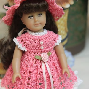 May include: A pink crocheted dress with white trim and a pink ribbon. The dress has a flower embellishment and is worn by a doll with brown hair and a pink crocheted hat.