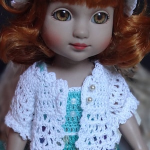 May include: A doll with red hair wearing a white crocheted cardigan over a teal and purple crocheted dress. The doll is wearing a white crocheted hat with purple flowers.