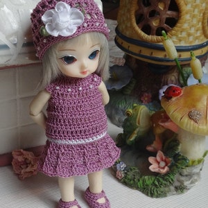 May include: A small doll wearing a purple crocheted dress and hat with a white flower. The doll is standing on a white surface with a miniature mushroom and a frog figurine.