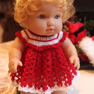 May include: A doll with blonde curly hair wearing a handmade red crochet dress with white trim. The doll also has matching red crochet shoes. The dress has a detailed openwork design.