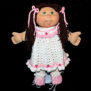 May include: A Cabbage Patch Kid doll wearing a white crocheted dress with pink trim and pink crocheted shoes. The doll has brown hair and green eyes.