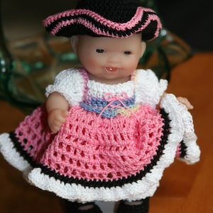 May include: A small doll wearing a handmade crochet outfit. The doll has a black hat with pink trim, a white top, and a pink skirt with black and white edging. The doll is wearing black shoes.