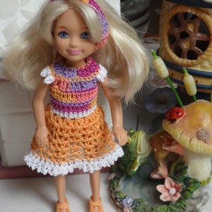 May include: A blonde doll wearing a crocheted dress with a pink, purple and orange striped bodice and a white crocheted trim. The doll is wearing a pink and white headband with a pearl accent. The doll is standing in front of a miniature mushroom and frog figurine.