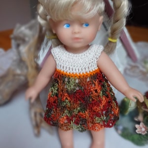 May include: A small doll wearing a crocheted dress with a white top and a multi-colored bottom. The dress has a brown, orange, green, and red pattern.