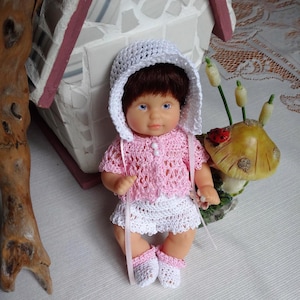 Puede incluir: Una muñeca pequeña con un vestido de crochet rosa y un gorro de crochet blanco. La muñeca tiene el pelo castaño y los ojos azules.