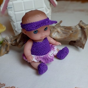 May include: A small purple crocheted dress with a white and pink ruffle trim, a purple crocheted hat with a visor, and purple crocheted booties. The doll is wearing the outfit.