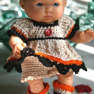 May include: A small doll wearing a crocheted dress with orange and black trim. The doll is wearing a black headband with a pumpkin-shaped embellishment. The doll is surrounded by small pumpkins.